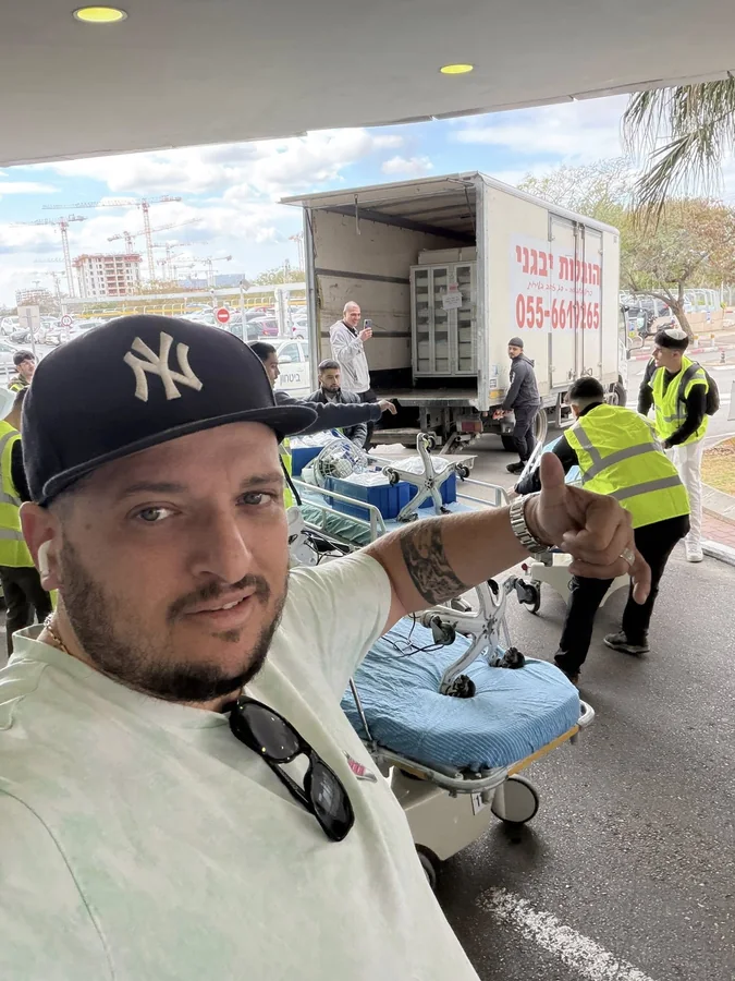 עובד חברת הובלות מצלם סלפי בזמן שצוות פורק מיטות אשפוז ממשאית הובלה לבנה.