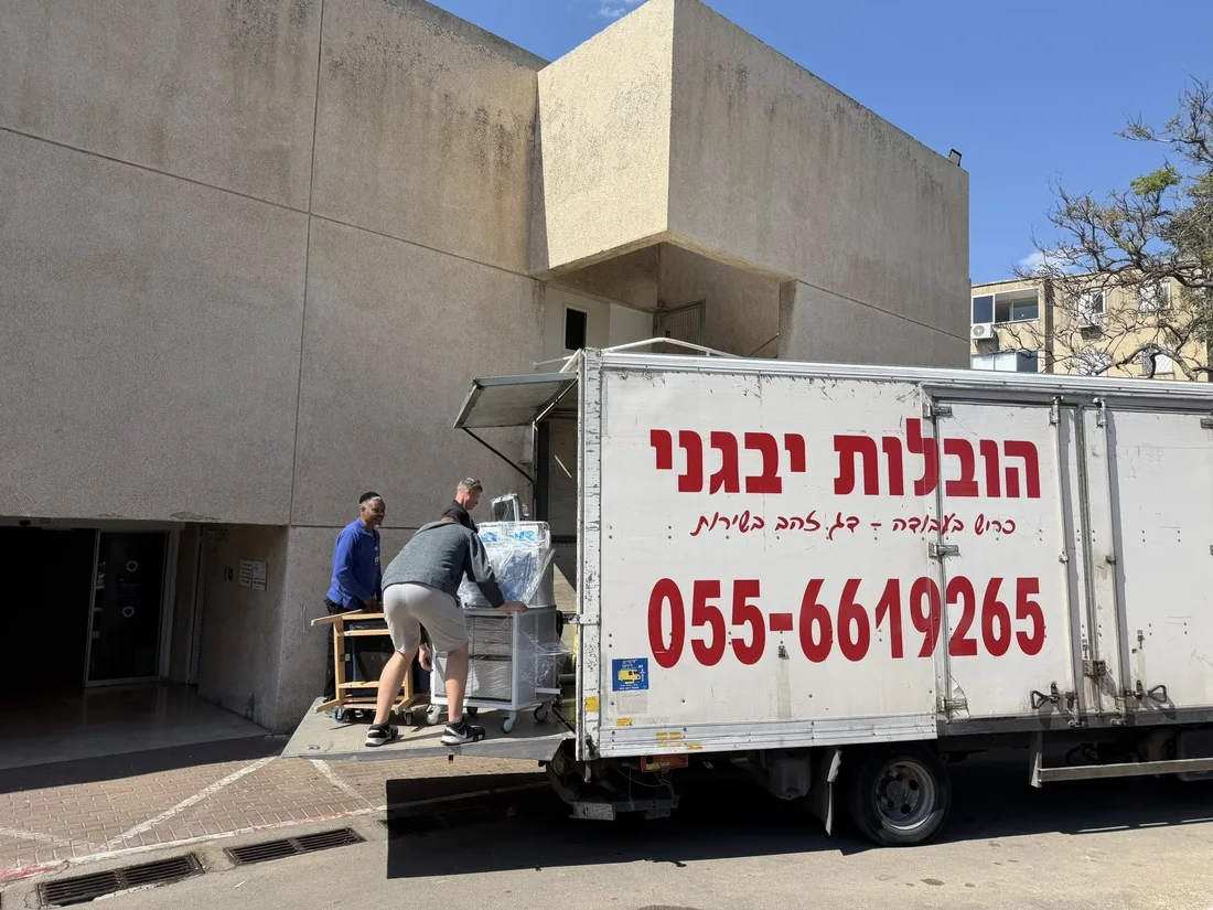 שני עובדי הובלות מעמיסים ציוד ארוז על עגלה למשאית לבנה עם כיתוב אדום.